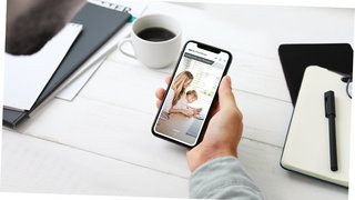 Schlotterer Magazin Hand hält Mobiltelefon in der Hand mit Kaffeetasse und Schreibunterlagen im Hintergrund