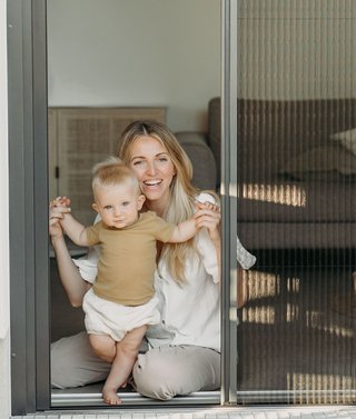 Blonde Frau mit Kind öffnet Insektenschutz Plissee Türe 