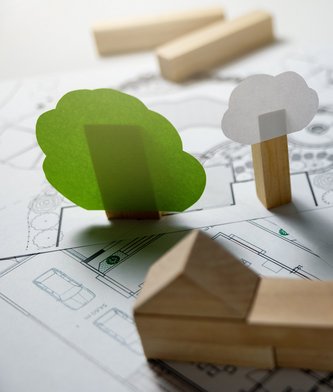 Ein nachhaltiges Haus wird auf einer gedruckten Planung durch stapelbare Holzsteinchen symbolisiert.