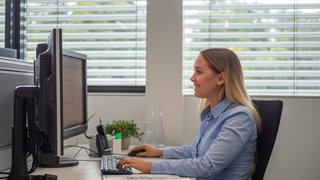 Frau sitzt am Schreibtisch und arbeitet am PC, im Hintergrund ist Sonnenschutz zu sehen.