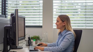 Sonnenschutz Arbeitsplatz Frau sitzt am Schreibtisch und arbeitet am PC, im Hintergrund ist Sonnenschutz zu sehen.